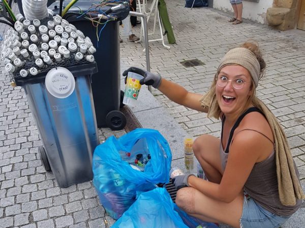 Das Foto zeigt Maxi, die einen Pfandbecher aus dem Müllsack gezogen hat und freudestrahlend nach oben hält beim Rudolstadt-Festival. Eine Aktion des Freie Bühne Jena e.V. und dem Projekt Theater WAGEN!