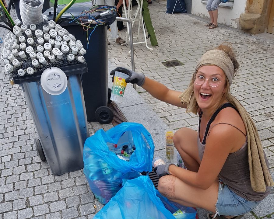 Das Foto zeigt Maxi, die einen Pfandbecher aus dem Müllsack gezogen hat und freudestrahlend nach oben hält beim Rudolstadt-Festival. Eine Aktion des Freie Bühne Jena e.V. und dem Projekt Theater WAGEN!